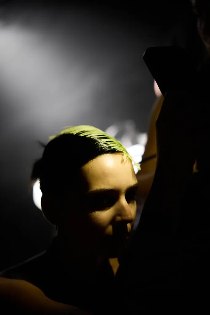 Couleur, en coulisses : le visage d’une personne aux cheveux teints en vert acide, pris dans une lumière rasante sur fond noir.