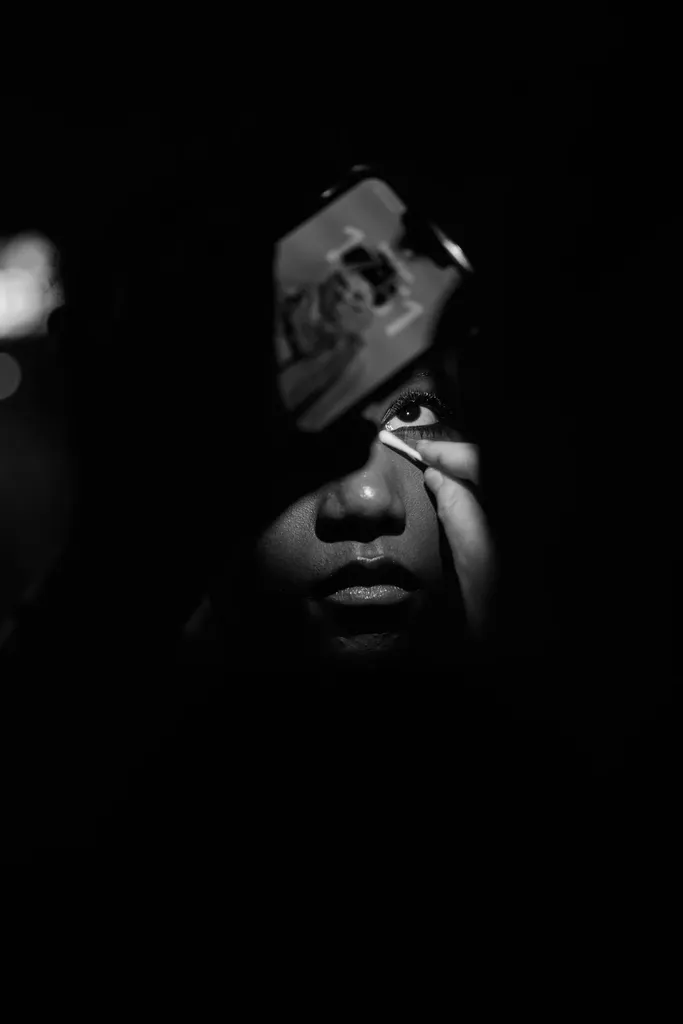 Noir et blanc, en coulisses : un visage dans l’ombre, un œil éclairé, pendant qu’une main applique le maquillage.