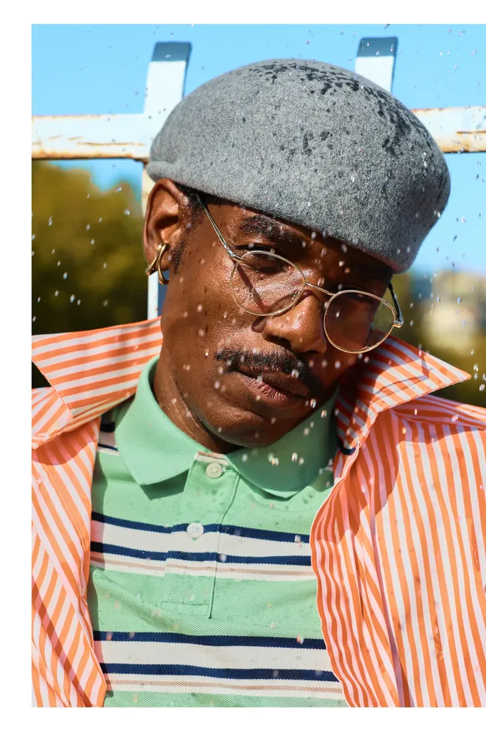 Couleur, gros plan en plein air : un homme en béret gris et lunettes rondes, chemise rayée orange sur polo vert, sous des gouttes d’eau.