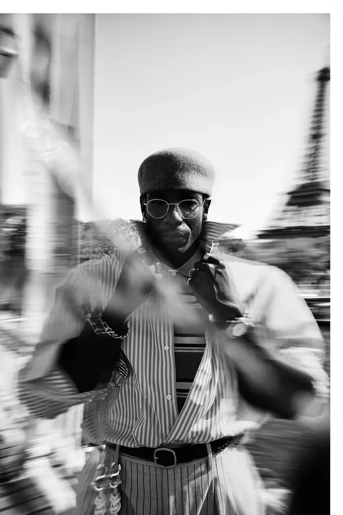 Noir et blanc, mouvement flou : un homme en béret et lunettes rondes, chemise rayée, ajuste son col dans la rue.