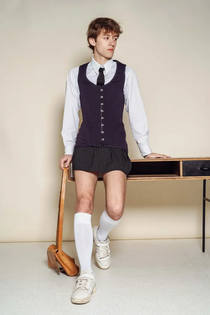 Couleur, en studio sur fond beige : un jeune homme en chemise blanche, gilet sombre, short et chaussettes hautes, appuyé contre un bureau.