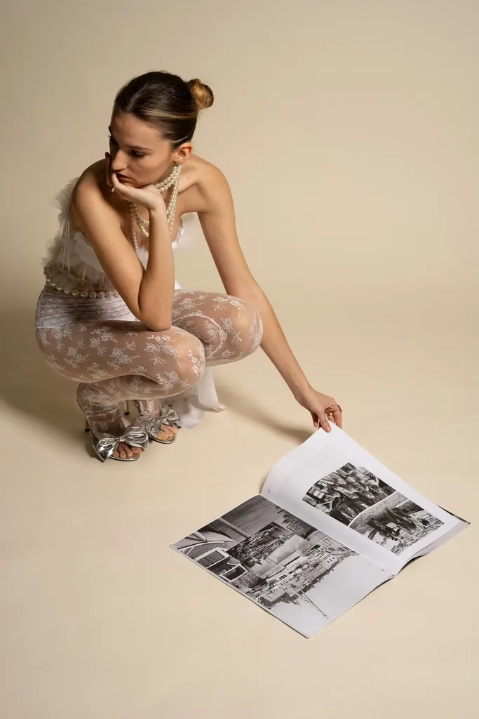 Couleur, en studio sur fond beige : une jeune femme accroupie en dentelle claire tourne les pages d’un magazine posé au sol.