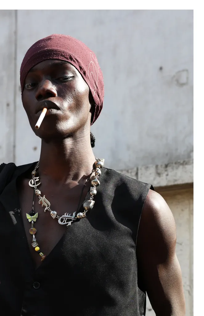 Couleur, portrait en plein jour : un homme en bandeau bordeaux et gilet noir, cigarette aux lèvres, colliers de perles et de breloques.