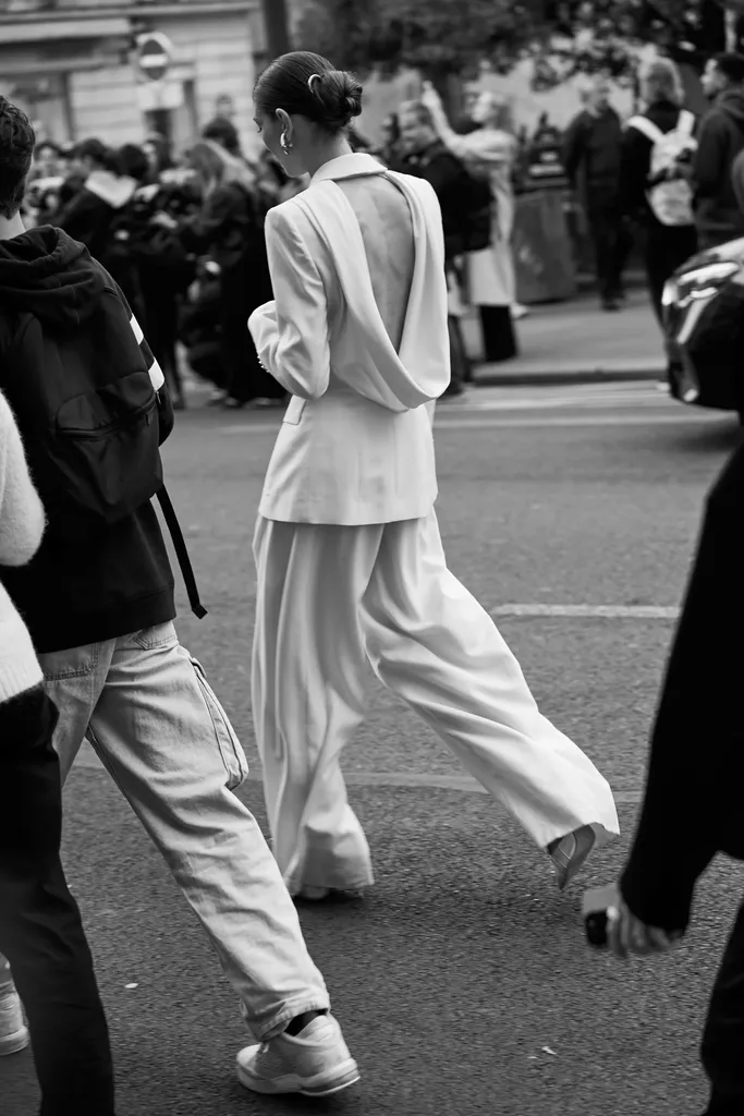 Noir et blanc, dans la rue : de dos, une femme en tailleur pantalon clair au dos ouvert traverse au milieu des passants.