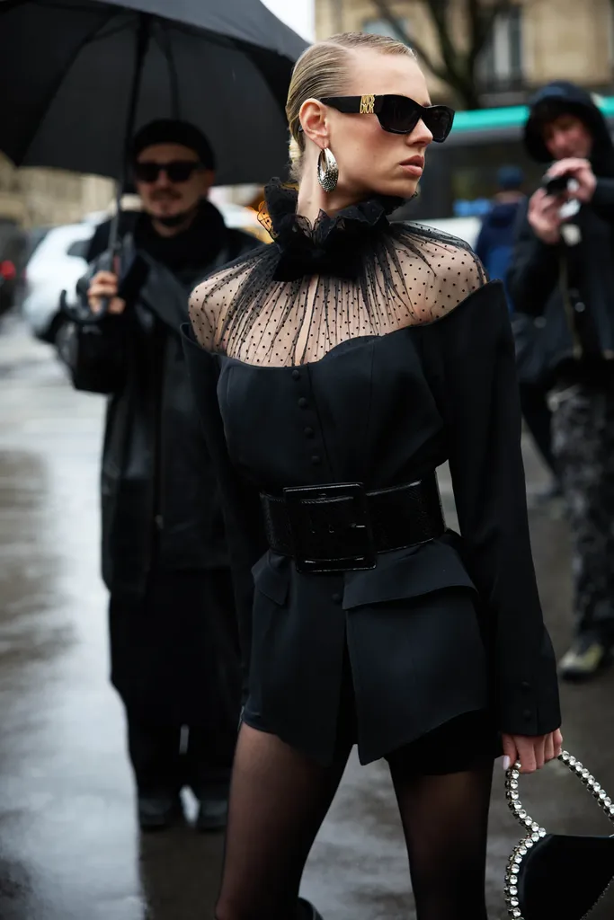 Couleur, sous la pluie : une femme en lunettes noires, col de tulle à pois et veste ceinturée, entourée de photographes.