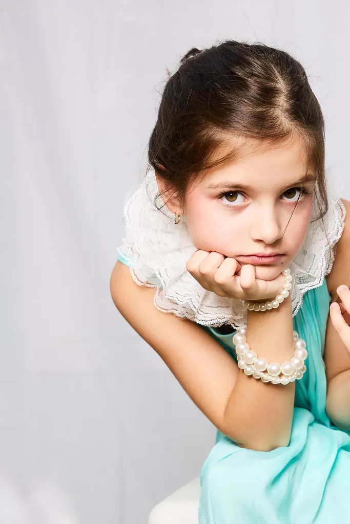 Fillette accoudée, le menton dans la main, col en dentelle, bracelets de perles et robe turquoise, sur fond clair.