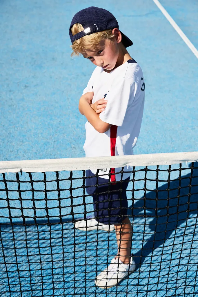 Garçon en casquette et tee-shirt blanc près du filet d’un court de tennis bleu, la tête baissée, en couleur.