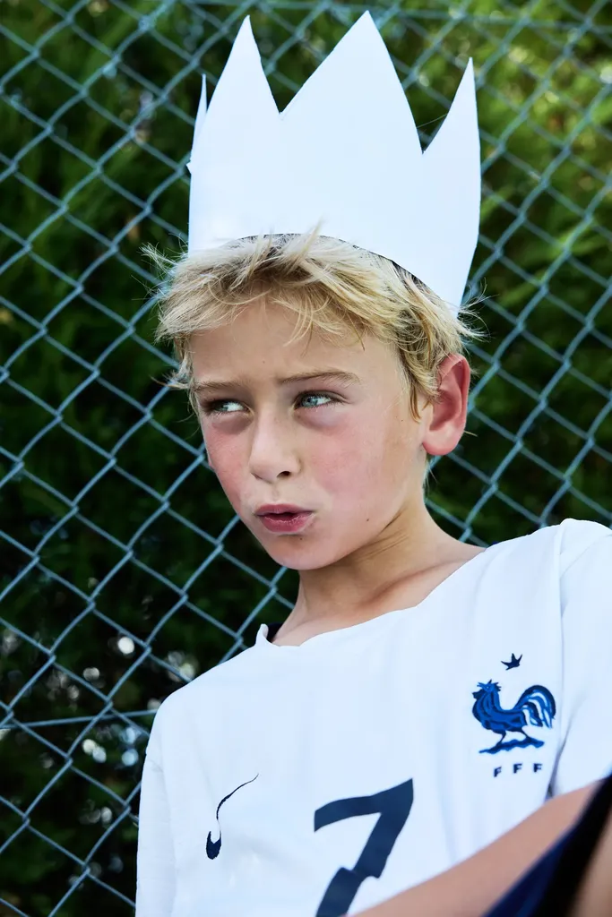 Garçon blond en couronne de papier et maillot blanc, le regard de côté, devant un grillage et des arbres.