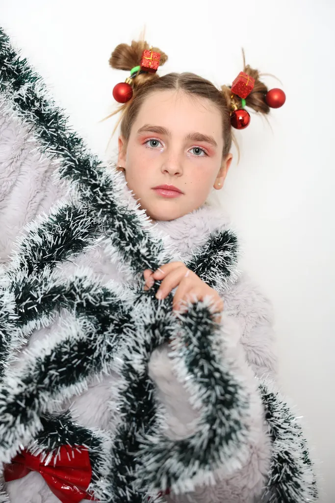 Jeune fille coiffée de deux chignons ornés de boules de Noël, enveloppée de guirlandes argentées et vertes, sur fond blanc.