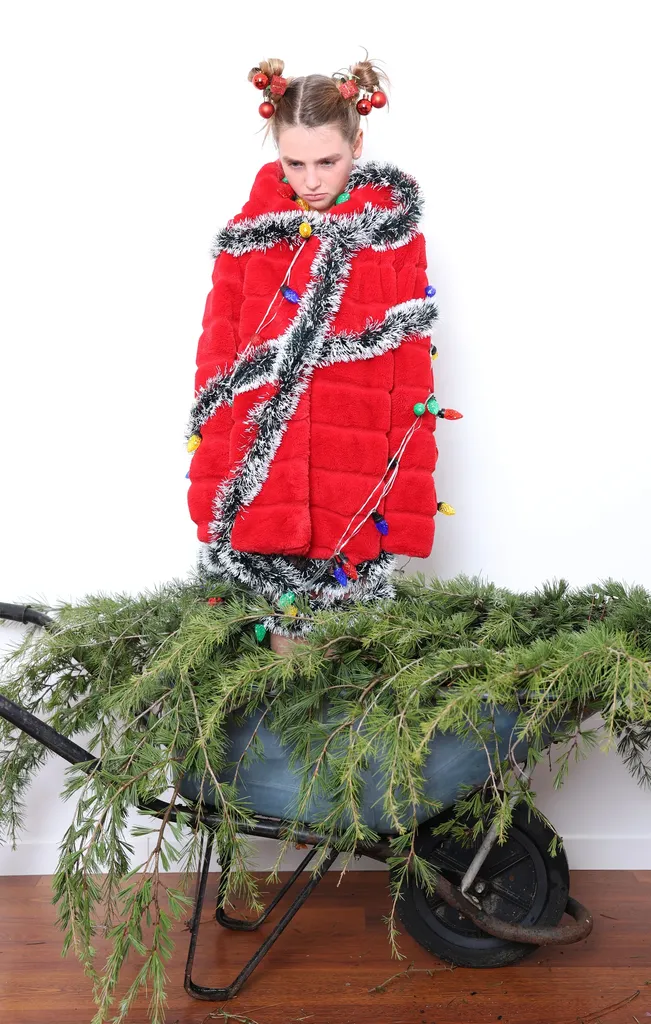 Jeune fille debout dans une brouette remplie de branches de sapin, manteau rouge enroulé de guirlande argentée, sur parquet.