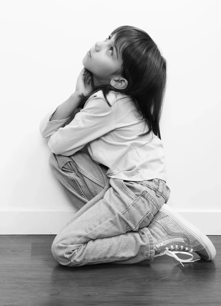 Fillette agenouillée contre un mur blanc, le visage levé, sur parquet, en noir et blanc.
