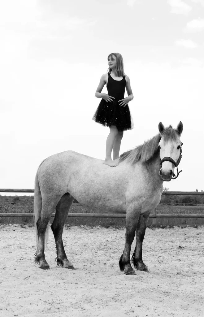 Jeune fille en tutu noir debout sur le dos d’un poney, dans un manège en sable, en noir et blanc.