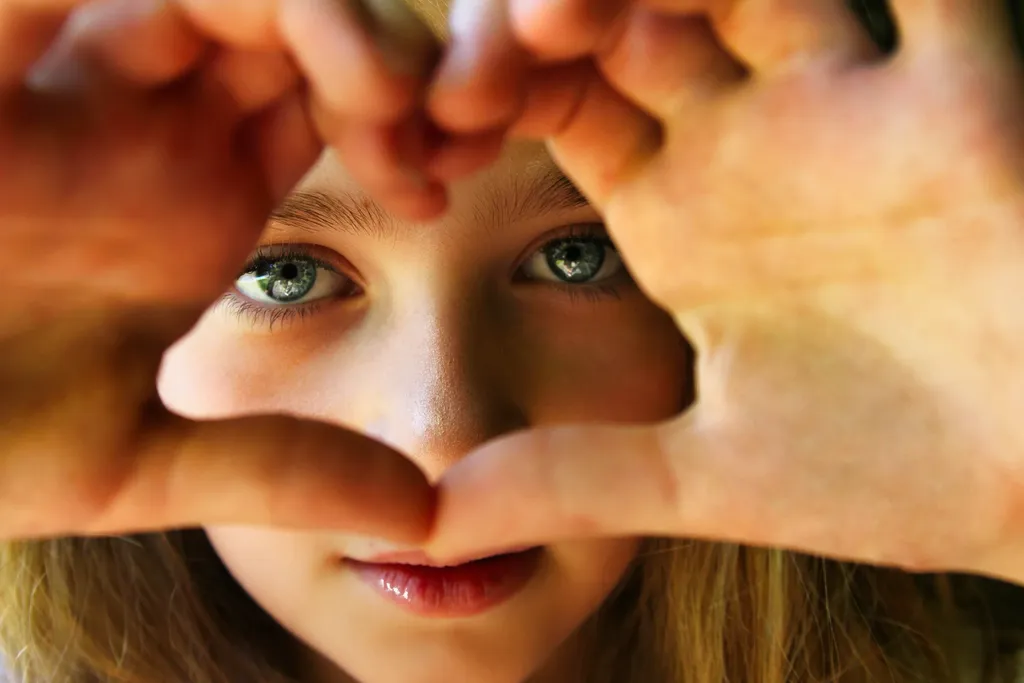 Gros plan sur des yeux verts encadrés par des mains formant un cœur, en couleur, lumière chaude.