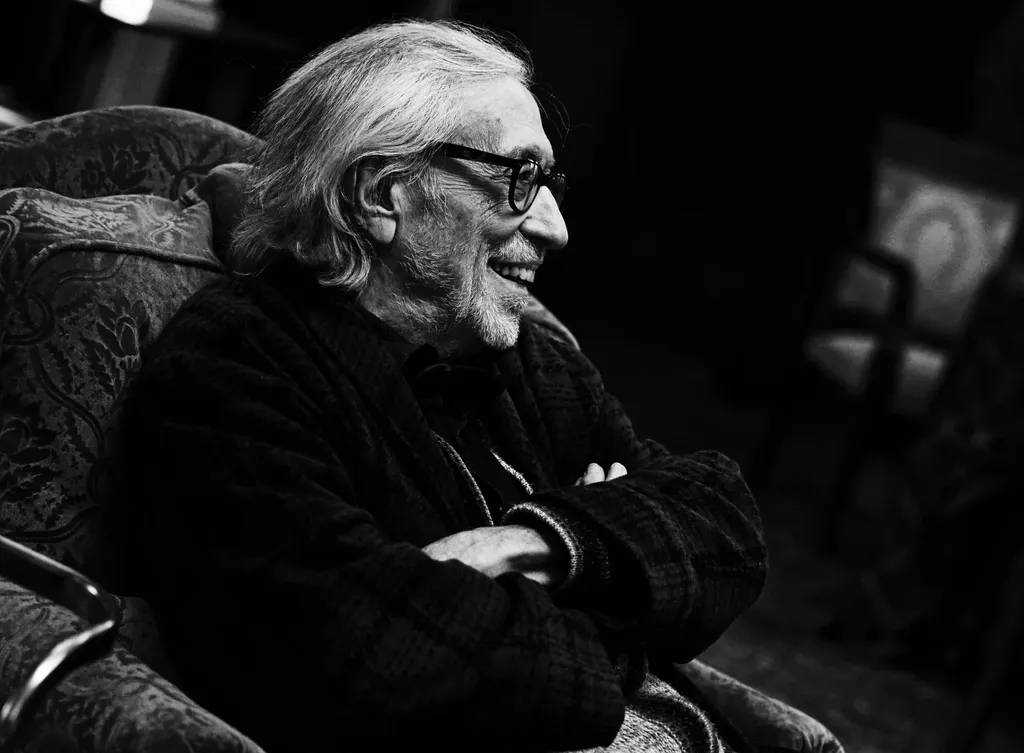 Black and white landscape portrait: an older man with white hair and glasses, smiling, arms folded in a patterned armchair.