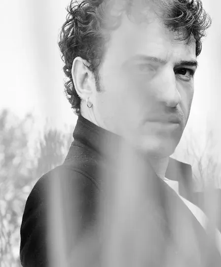 Black and white portrait of a man with curly hair, an earring and a high-collared coat, seen through a blurred foreground.