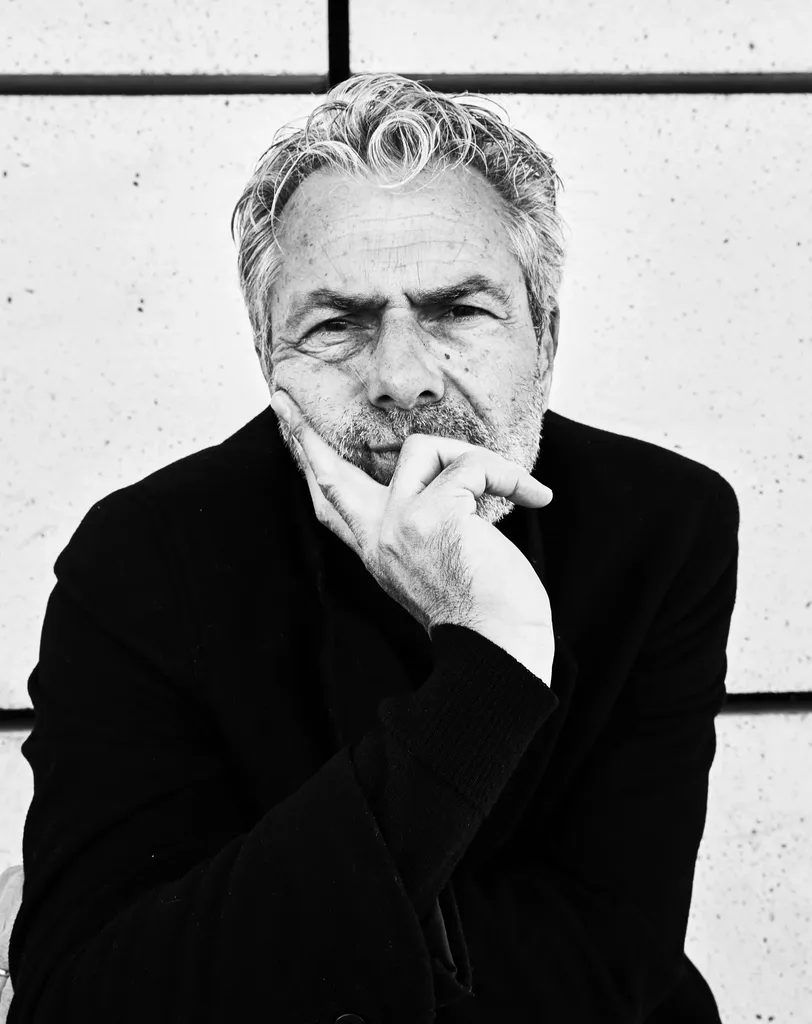 Black and white portrait of a grey-haired man, hand under his chin, in a dark coat against a pale wall.